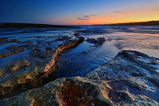 Sunrise seascape with orange rocks and ocean pools and sea shells with clean bright sky and distant city lights