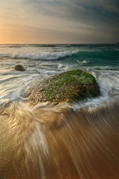 Sunrise seascape with a lone mossy rock with a flowing wave around it