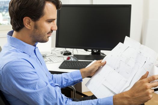 Young handsome man sitting at his desk in the office while reading written agreements and studying important documentation for work