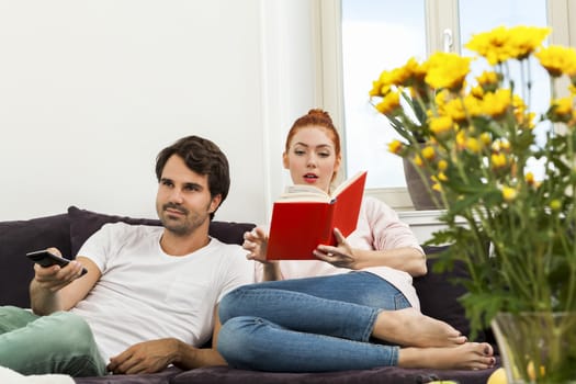 Young Couple Resting on the Sofa at the Living Room While the Man is Watching TV and Woman is Reading a Book.