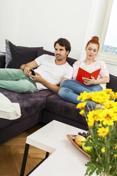 Young Couple Resting on the Sofa at the Living Room While the Man is Watching TV and Woman is Reading a Book.