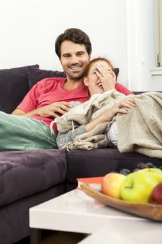 Young Couple Resting on the Sofa at the Living Room While the Man is Watching TV and Woman is Reading a Book.