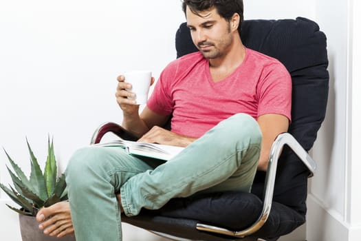 Young Man in Casual Clothing Sitting on Black Chair While Reading a Book and Holding a Glass of Drink.