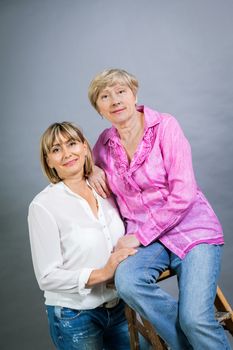 Attractive stylish blond senior lady with her beautiful middle-aged daughter posing together with her hands on her shoulders smiling at the camera on a grey studio background