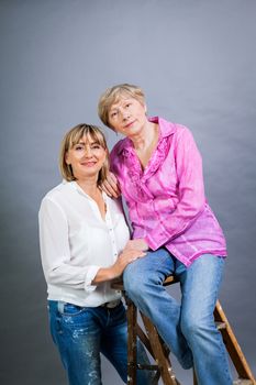 Attractive stylish blond senior lady with her beautiful middle-aged daughter posing together with her hands on her shoulders smiling at the camera on a grey studio background