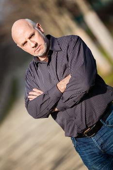 Confident attractive middle-aged man standing waiting in a rural lane with folded arms looking to the right hand side of the frame as though expecting somebody to arrive, with copyspace