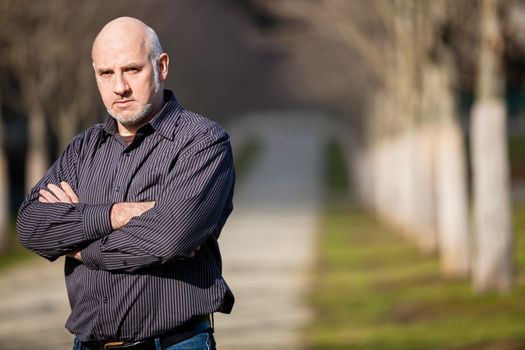 Confident attractive middle-aged man standing waiting in a rural lane with folded arms looking to the right hand side of the frame as though expecting somebody to arrive, with copyspace