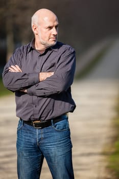 Confident attractive middle-aged man standing waiting in a rural lane with folded arms looking to the right hand side of the frame as though expecting somebody to arrive, with copyspace