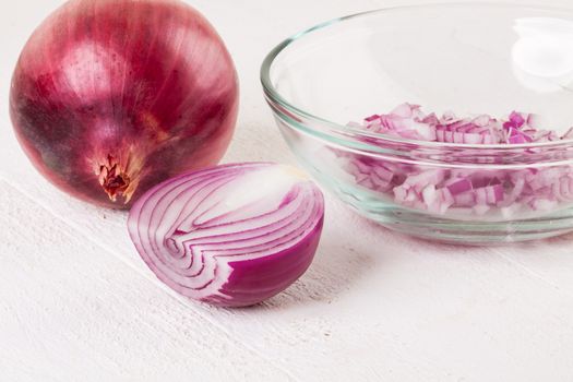 Heap of fresh finely diced red onion with a peeled half showing a cross-section and whole unpeeled one behind on a white background