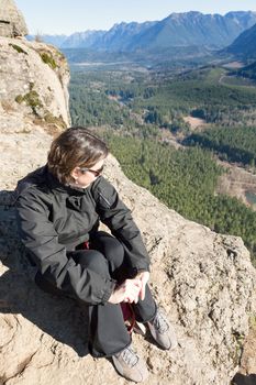 Young woman wearing sunglasses sitting on a rocky outcrop enjoying the view of the forested valley, lake and mountains from Rattlesnake Ledge Trail, Snoqualmie, Washington after hiking up the summit