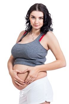 Young pregnant woman making a heart gesture with her hands against her bare belly showing her love of her unborn child while looking at the camera with a smile, isolated on white