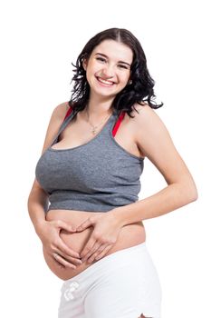 Young pregnant woman making a heart gesture with her hands against her bare belly showing her love of her unborn child while looking at the camera with a smile, isolated on white