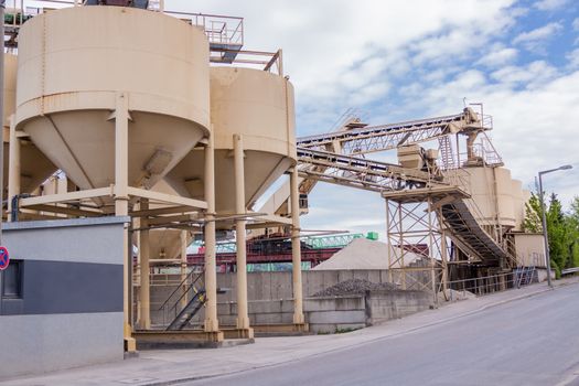 Row of large cylindrical metal tanks or silos at a refinery plant , mill or factory with an infrastructure of conveyor belts against a cloudy blue sky