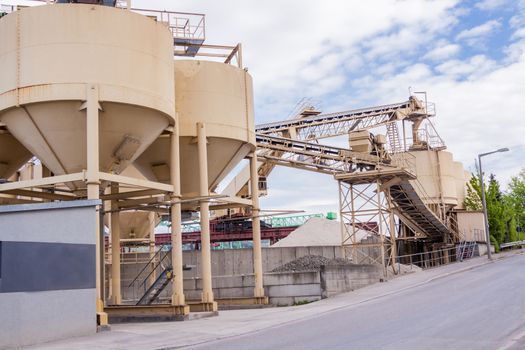 Row of large cylindrical metal tanks or silos at a refinery plant , mill or factory with an infrastructure of conveyor belts against a cloudy blue sky