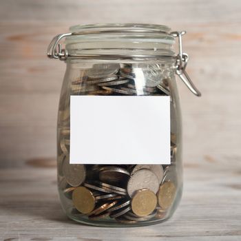 Coins in glass money jar with white blank label, financial concept. Vintage wooden background with dramatic light.