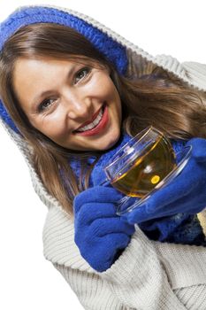 Fashionable young woman in a blue knitted winter ensemble and cowl neck jersey sipping a cup of hot tea with a smile in an effort to keep warm, isolated on white