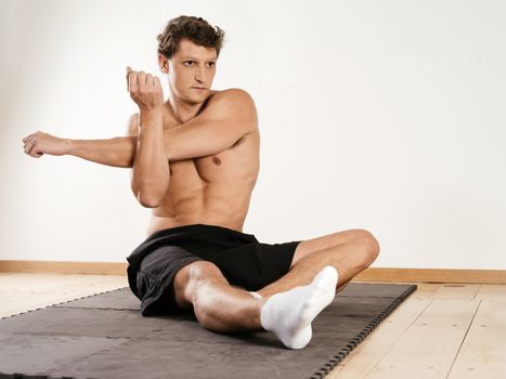 Photo of a young attractuive man stretching his shoulder muscles while sitting on exercise mat.
