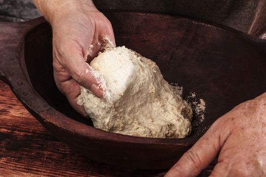 Old female hands knead whole wheat dough in vintage wooden bowl on wooden background. Traditional healthy baking, rustic vintage style.