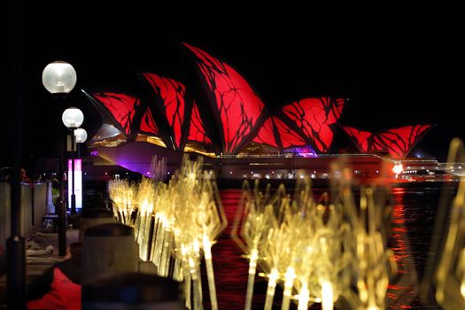SYDNEY, AUSTRALIA - MAY 27, 2015;  The Sydney Opera House in vivid red and harbour reflections.  In the foreground another exhbit entitled Tidal Reed Garden leads the viewers eye along the wharf towards the Opera House. Focus is to the Opera House.