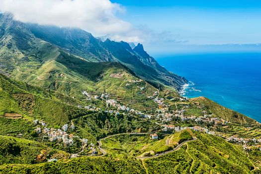 Small city or village buildings and winding or serpantine road in green mountain or rock valley and clouds with horizon landscape near coast of Atlantic ocean in Tenerife Canary island, Spain