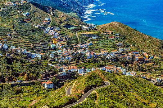 Small village buildings and winding or serpantine road in green mountain valley landscape near the coast or shore of Atlantic ocean in Tenerife Canary island, Spain