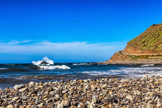 Wild stone beach and mountain on coast or shore of the Atlantic ocean with waves and sky with clouds and skyline or horizon in Tenerife Canary island, Spain at spring or summer