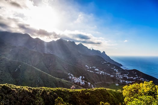 Green mountains or rock with clouds and sun with sunlight and sunbeams on sky over small village or city near the Atlantic ocean in Tenerife Canary island, Spain
