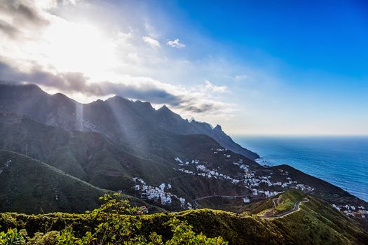 Coast or shore of Atlantic ocean with green mountain or rock and blue sky with clouds and sun with sunlight and horizon in Tenerife Canary island, Spain at spring or summer