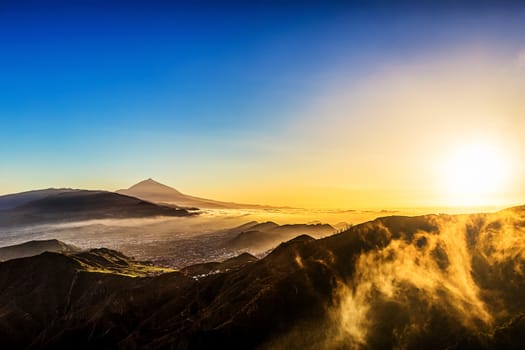 Sun with sunlight over mountains on blue sky and clouds with haze and Teide volcano on background at evening sunset in Tenerife Canary island, Spain