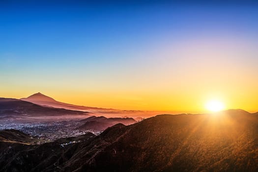 Sun with sunlight over mountains on blue sky with fog and Teide volcano on background at evening sunset in Tenerife Canary island, Spain at spring or summer