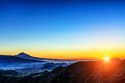 Sun over mountains on blue sky with fog or haze and Teide volcano on background at evening sunset in Tenerife Canary island, Spain at spring or summer