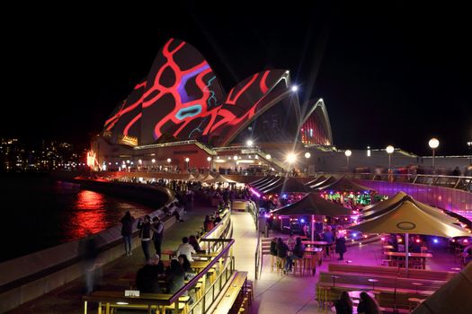 SYDNEY, AUSTRALIA - MAY 25, 2015;   Sydney Opera House roof lit up with various colourful patterns during Vivid Sydney annual festival event. Tourists and locals dine and relax in the Opera Bar in foreground and other restaurants along the forecourt