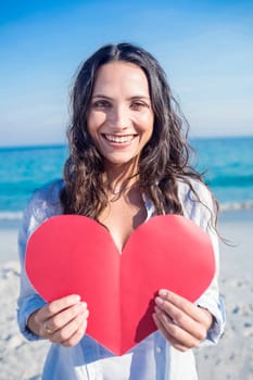 Smiling woman holding heart card at the beach on a sunny day