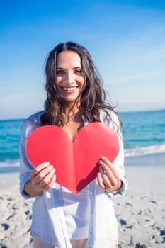 Smiling woman holding heart card at the beach on a sunny day