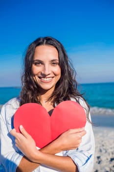 Smiling woman holding heart card at the beach on a sunny day