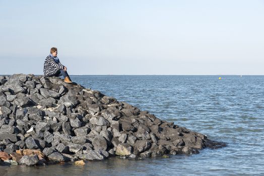 adult white woman with brown boots and blue clothes sitting on the rocks near the sea and looking to the horizon waiting for something and thinking 