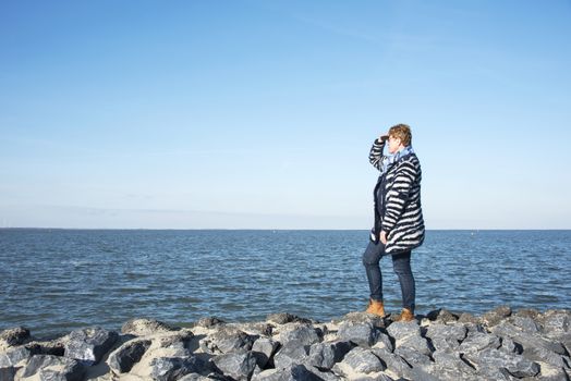 adult woman in blue clothes and brown boots standing on the rocks at the coast and staring to the horizon over the empty sea