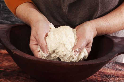 Old female hands knead whole wheat dough in vintage wooden bowl on wooden background. Traditional healthy baking, rustic vintage style.