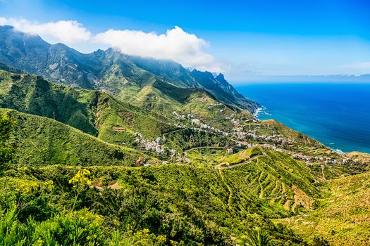 Small city or village buildings and winding or serpantine road in green mountain or rock valley and clouds with horizon landscape near coast or shore of Atlantic ocean in Tenerife Canary island, Spain