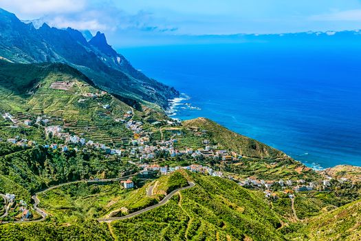 Small city or village buildings and winding or serpantine road in green mountain or rock valley and clouds with horizon landscape near shore of Atlantic ocean in Tenerife Canary island, Spain