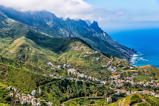 City or village buildings and winding or serpantine road in green mountain or rock valley and clouds with horizon landscape near coast of Atlantic ocean in Tenerife Canary island, Spain