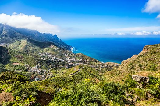 Small village buildings and winding road in green mountain or rock valley and clouds with horizon landscape near coast or shore of Atlantic ocean in Tenerife Canary island, Spain