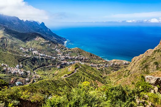 Village buildings and winding or serpantine road in green mountain or rock valley and clouds with horizon landscape near coast or shore of Atlantic ocean in Tenerife Canary island, Spain