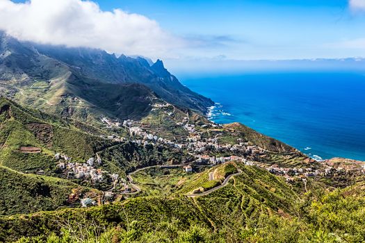 Small city or village and winding or serpantine road in green mountain or rock valley and clouds with horizon landscape near  coast of Atlantic ocean in Tenerife Canary island, Spain