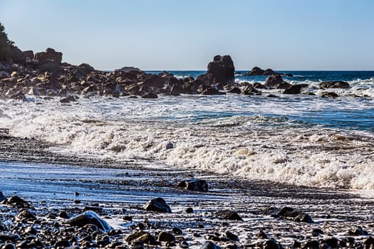 Waves and foam on wild stone beach on coast or shore of the Atlantic ocean with sky and rock on skyline or horizon in Tenerife Canary island, Spain