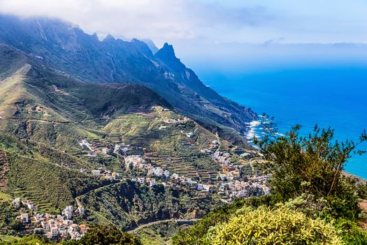 Village buildings and winding or serpantine road in green mountain or rock valley and clouds with horizon landscape near shore of Atlantic ocean in Tenerife Canary island, Spain