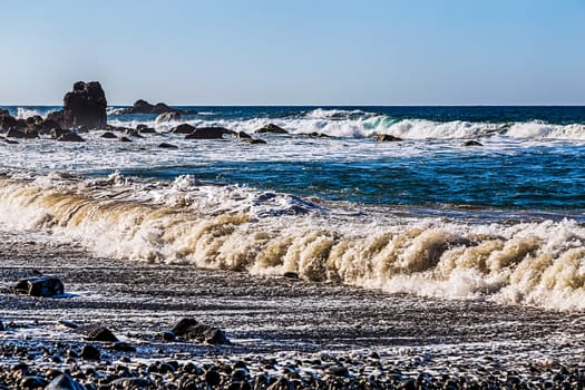 Waves and foam on wild stone beach on shore of the Atlantic ocean with sky and rock on skyline or horizon in Tenerife Canary island, Spain at spring or summer