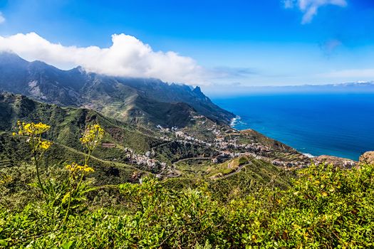 City or village and winding or serpantine road in green mountain or rock valley and clouds with horizon landscape near coast or shore of Atlantic ocean in Tenerife Canary island, Spain