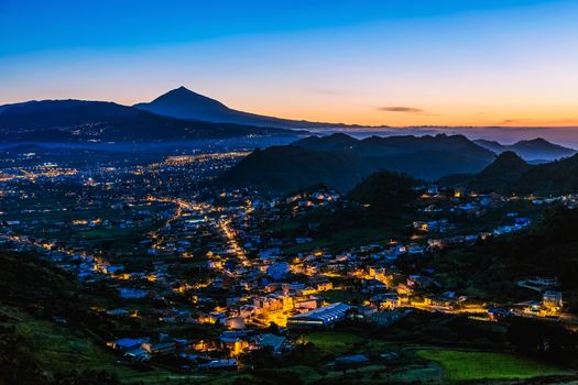 City or town with illumination after sunset or sundown at evening in mountains with blue sky and Teide volcano on background in Tenerife Canary island, Spain