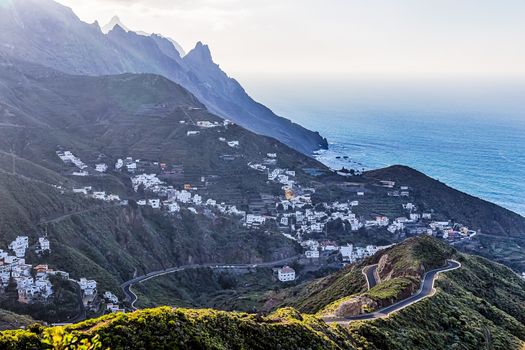 City or village and winding or serpantine road in green mountain or rock valley and clouds with horizon landscape near shore of Atlantic ocean in Tenerife Canary island, Spain